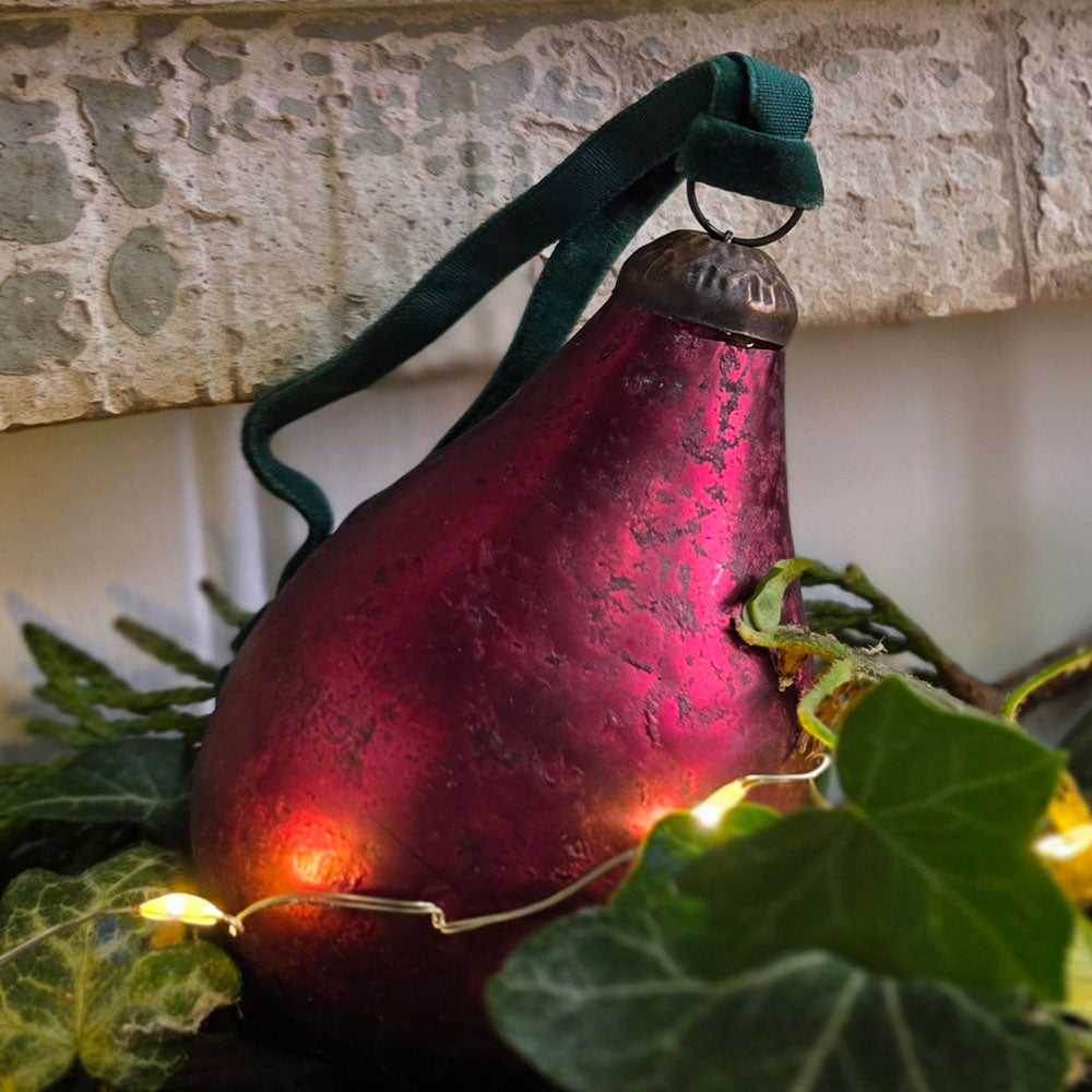 A textured, pear-shaped Christmas ornament in a deep burgundy red with a dark green velvet hanging ribbon, styled with green ivy leaves and warm fairy lights against a stone wall background.