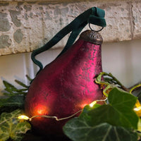 A textured, pear-shaped Christmas ornament in a deep burgundy red with a dark green velvet hanging ribbon, styled with green ivy leaves and warm fairy lights against a stone wall background.
