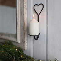A white pillar candle lit in a black metal wall sconce with a heart-shaped top, mounted on a white paneled wall next to a rustic mirror and evergreen garland.