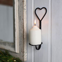 A black metal wall candle holder with a heart-shaped top and a lit white pillar candle, mounted on a light gray paneled wall next to a rustic mirror frame.