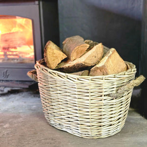 Pale willow wood basket filled with logs by a lit wood burning stove.