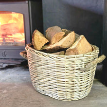 Oval willow wood basket filled with cut logs next to a lit wood burning stove.