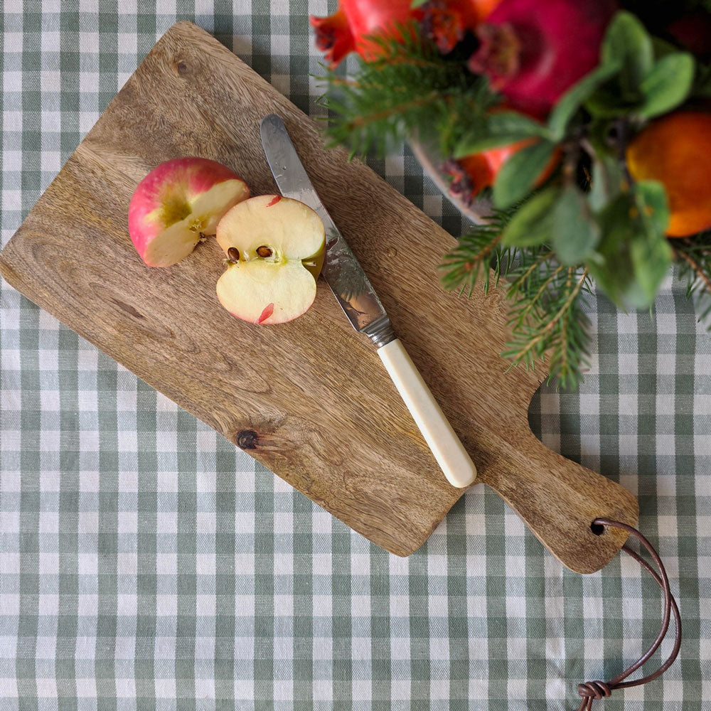 A rustic wooden cutting board with a handle, styled with a knife, sliced apples, and a floral arrangement on a green and white checkered tablecloth.