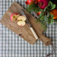 A rustic wooden cutting board with a handle, styled with a knife, sliced apples, and a floral arrangement on a green and white checkered tablecloth.