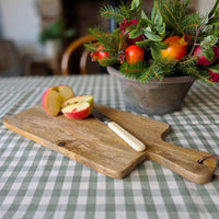 Small paddle shaped mango wood board on a green checked tablecloth with a chopped apple next to a terracotta bowl filled with foliage and pomegranate.