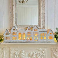 A decorative row of small house-shaped Christmas ornaments with glowing warm lights inside, displayed on a white mantelpiece in front of a mirror.