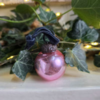 A pink glass Christmas ornament with a dark navy blue ribbon loop and a dark metal cap, resting on green ivy leaves with warm string lights in the background.