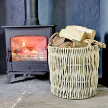 Round wicker Cotswold log basket on a stone hearth filled with with logs next to a lit wood burning stove.