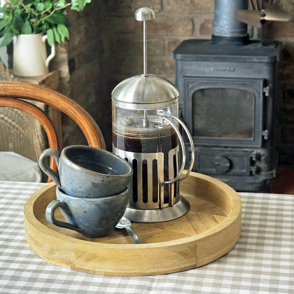 Coffee cafetiere and blue ceramic cups on a small circular wooden serving tray on a table covered with gingham tablecloth in front of a wood burning stove.