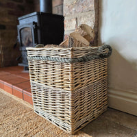 A natural wicker storage basket with a thick braided rope handle, filled with firewood logs, set on a carpeted hearth next to a brick fireplace and wood stove.
