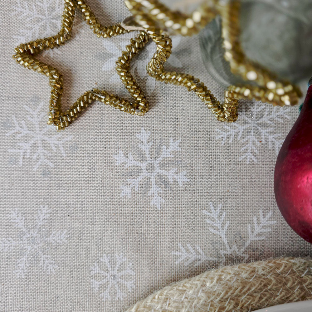 A close-up of a beige fabric surface with a white snowflake pattern, styled with a gold beaded star-shaped decoration, a red glass ornament, and a woven straw-like prop.