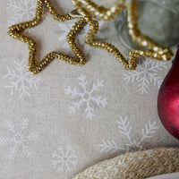 A close-up of a beige fabric surface with a white snowflake pattern, styled with a gold beaded star-shaped decoration, a red glass ornament, and a woven straw-like prop.