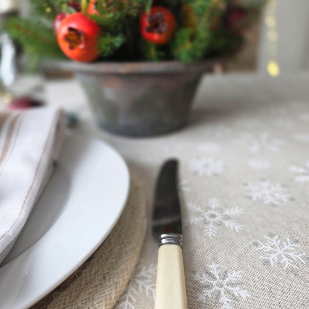 A close-up of a beige linen tablecloth printed with white snowflakes, styled with a white plate, folded cream napkin, and a butter knife with a cream handle. A rustic metal pot of holiday greenery and red berries sits blurred in the background.