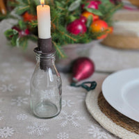 A lit white taper candle in a dark metal holder set inside a small clear glass bottle, styled on a snowflake-patterned tablecloth with holiday decorations in the background.