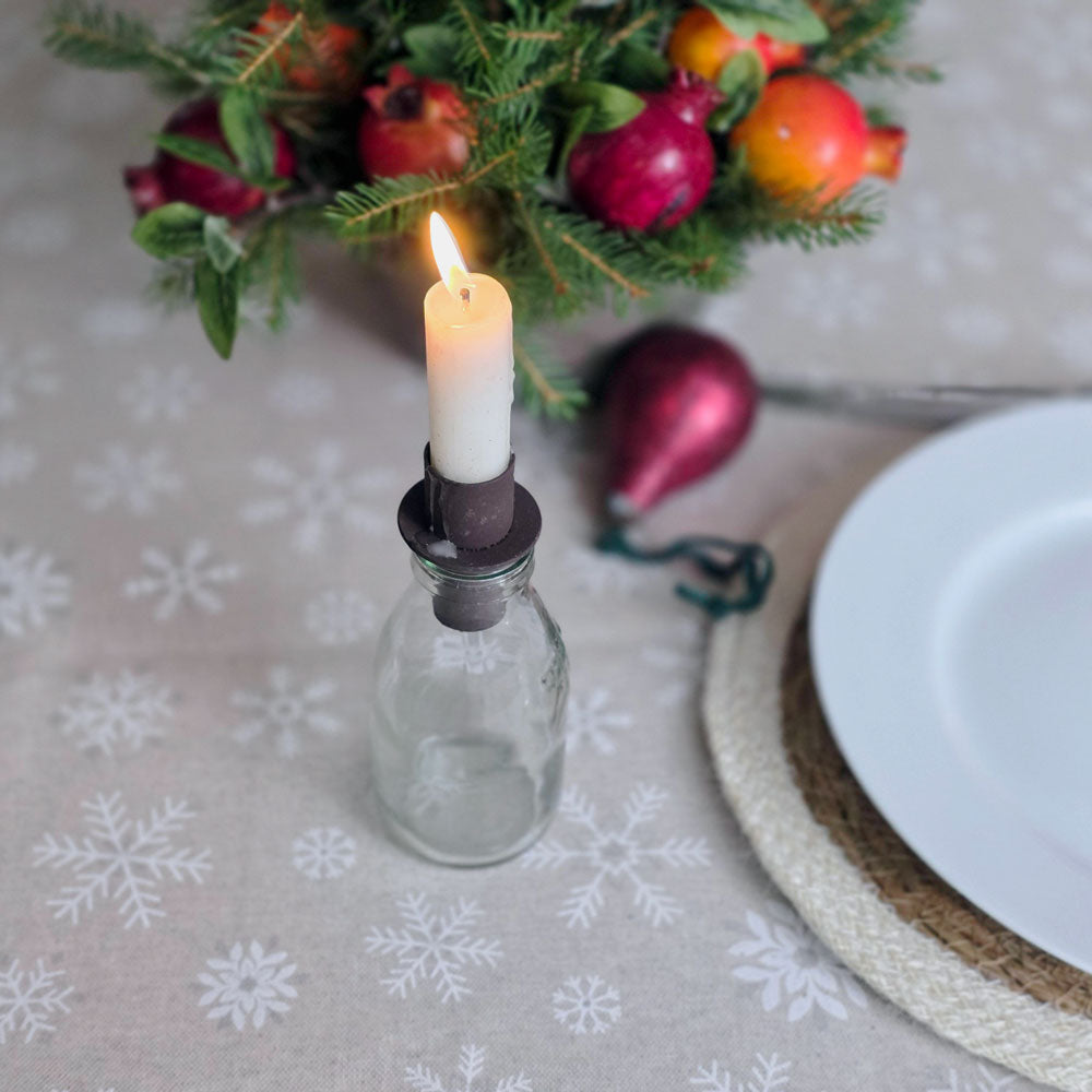 A lit white candle in a dark metal holder mounted on a small clear glass bottle, styled on a beige snowflake-patterned tablecloth with a white plate, woven placemat, and holiday greenery in the background.