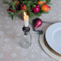 A lit white candle in a dark metal holder mounted on a small clear glass bottle, styled on a beige snowflake-patterned tablecloth with a white plate, woven placemat, and holiday greenery in the background.