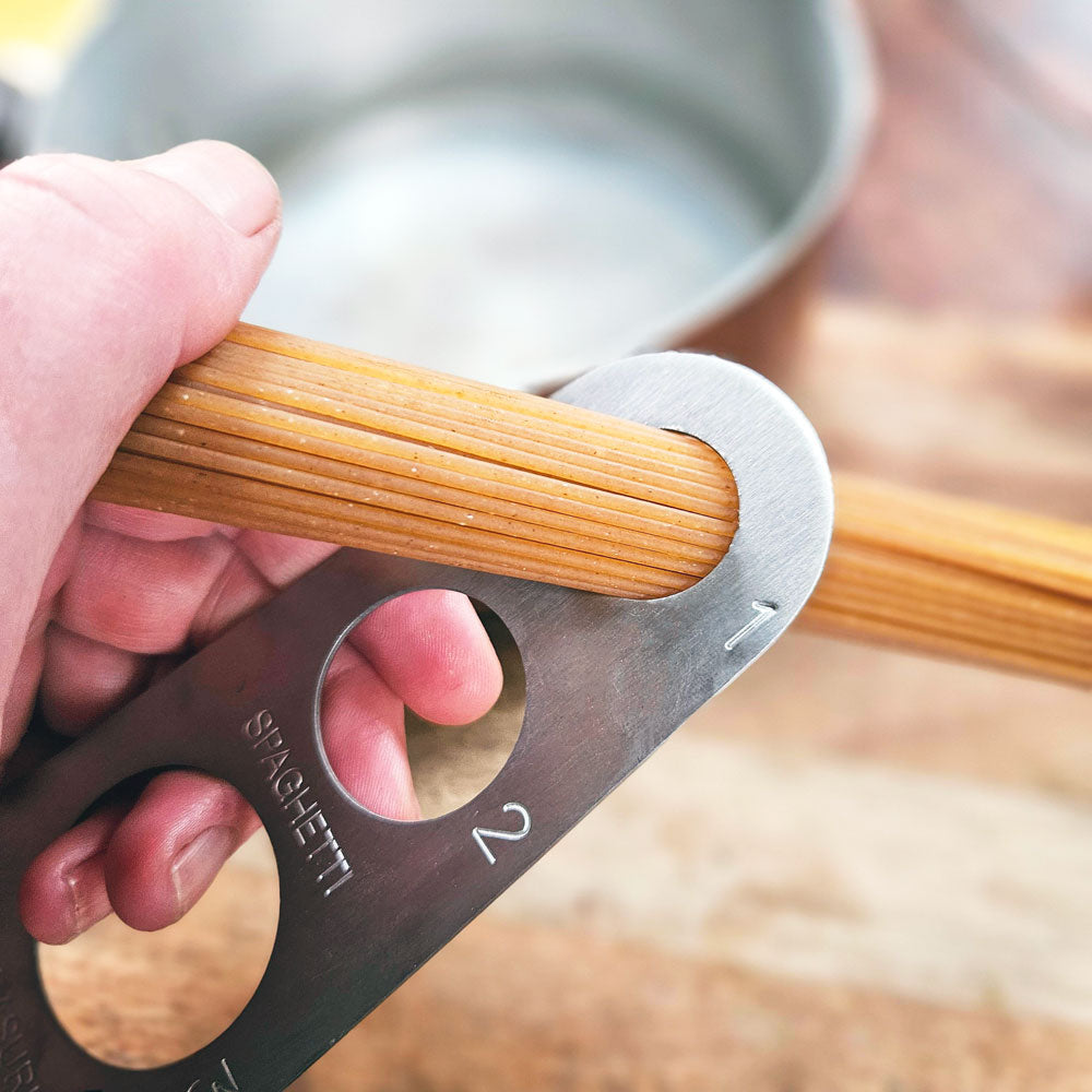 Hand holding a metal tool with cut-out shapes for measuring pasta, with a blurred background of a pot and noodles.