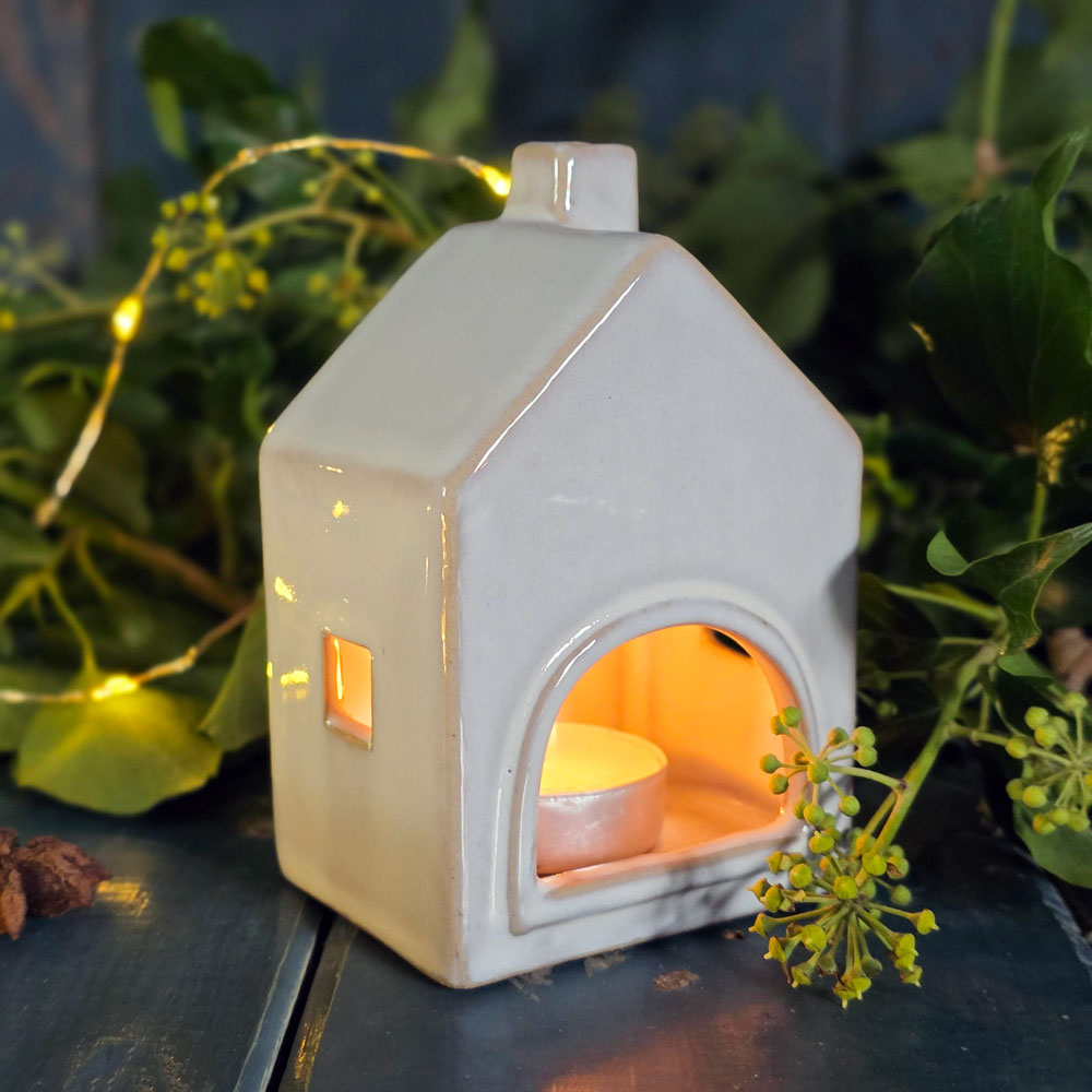 Rear view of ceramic house-shaped lantern with a small candle inside, surrounded by greenery.