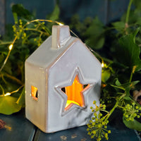 Ceramic house-shaped lantern with a star cutout, glowing inside, surrounded by greenery.