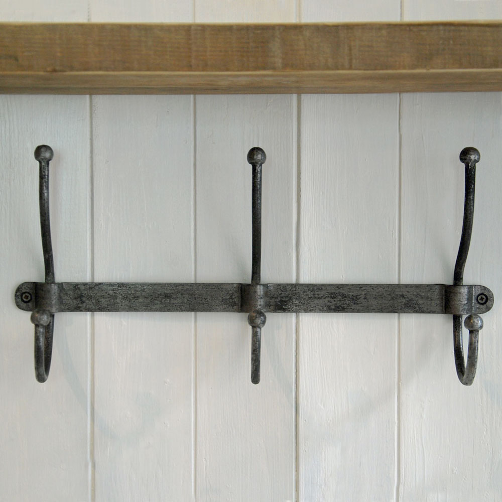 A rustic three-hook wall coat rack made of dark weathered metal, mounted on a white paneled wall with a wooden shelf above.