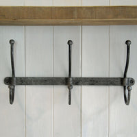 A rustic three-hook wall coat rack made of dark weathered metal, mounted on a white paneled wall with a wooden shelf above.