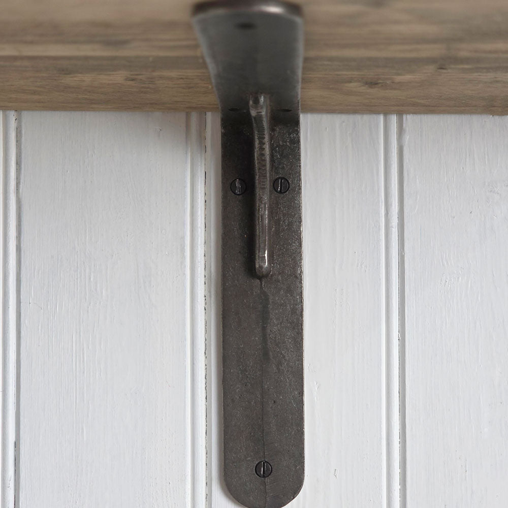 A rustic dark metal shelf bracket with a curved hook design, shown mounted against a white paneled wall with a light wood shelf above it.