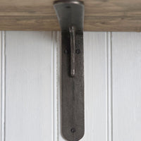 A rustic dark metal shelf bracket with a curved hook design, shown mounted against a white paneled wall with a light wood shelf above it.