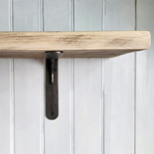 A close-up of a rustic light wood shelf mounted on a white vertical paneled wall with a dark metal bracket support.