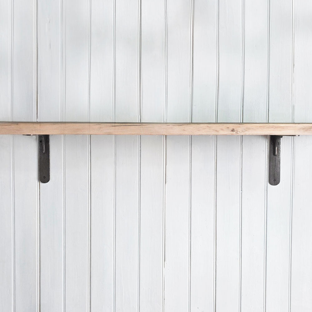 A rustic light wood shelf mounted horizontally on a white vertical paneled wall, supported by two dark metal brackets.