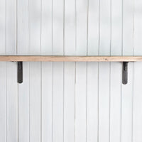 A rustic light wood shelf mounted horizontally on a white vertical paneled wall, supported by two dark metal brackets.