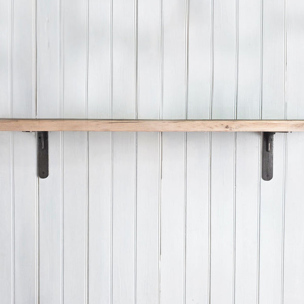 A rustic light wood shelf mounted horizontally on a white vertical paneled wall, supported by two dark metal brackets.