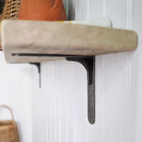 A rustic dark metal L-bracket shelf mount holding a light beige shelf with a terracotta pot on a white paneled wall.