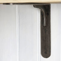 A dark metal L-bracket shelf mount shown attached to a white paneled wall, with part of a light wood shelf edge visible at the top.