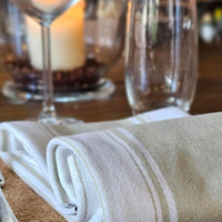 A folded white linen napkin with subtle horizontal stripes, styled on a wooden table next to a wine glass and a blurred candle.