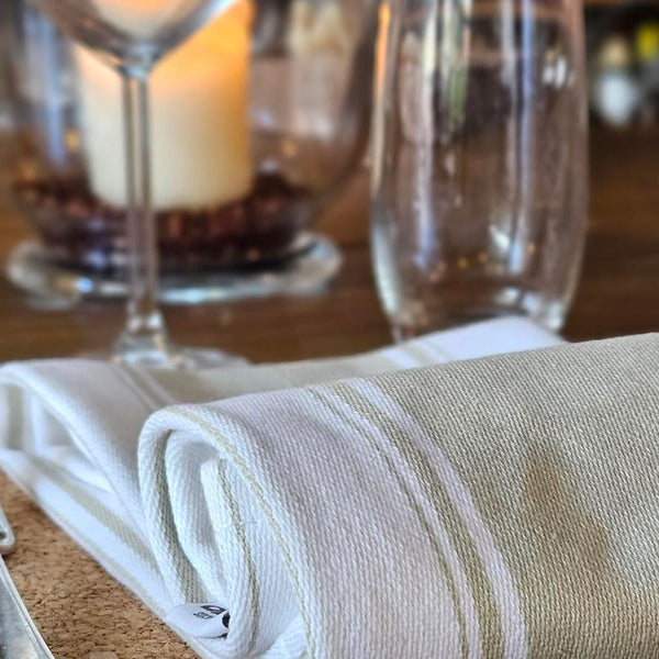 A folded white linen napkin with subtle horizontal stripes, styled on a wooden table next to a wine glass and a blurred candle.