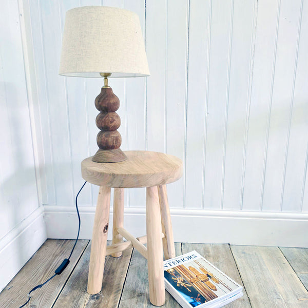 Wooden stool with a lamp on top against a white paneled wall.
