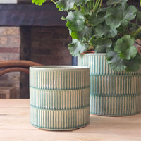 Two ribbed ceramic planters in a muted sage green glaze, with one in the foreground on a wooden table and another behind it holding a green leafy plant, set against a softly blurred indoor background.