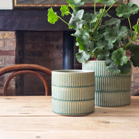 Two green glazed indoor plant pots on a scrubbed kitchen table in front of stove and chair. Large Pelargonium planted in the larger pot.