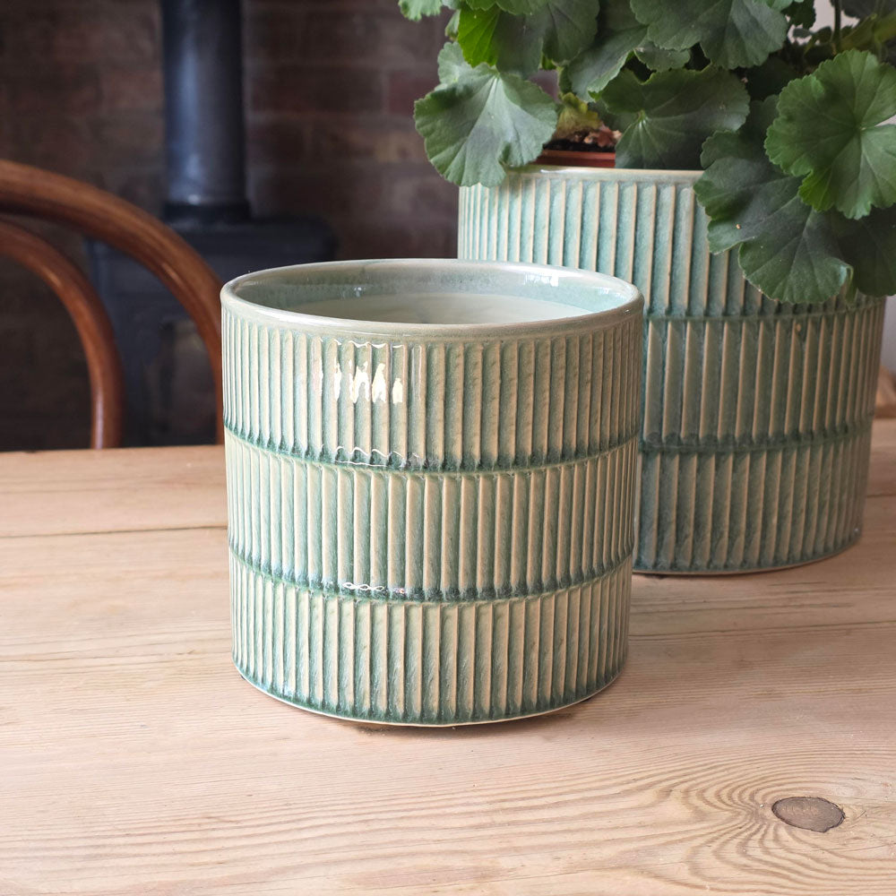 A ribbed ceramic planter in a sage green glaze sits on a light wood table, with a second matching planter and green foliage in the background.