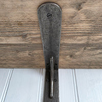 A rustic metal shelf bracket with a distressed dark gray finish, shown mounted against a wooden beam and white paneled wall.