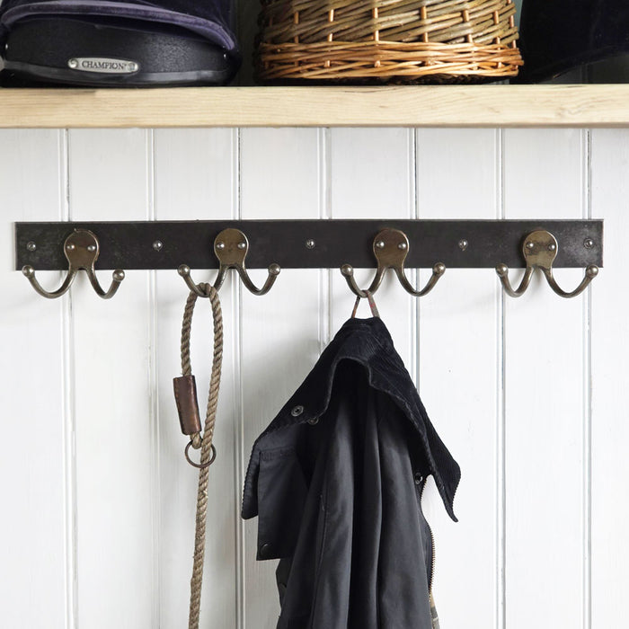 A dark metal wall hook rack with five hooks mounted on a white paneled wall, with a black coat hanging from one hook and a rope bag hanging from another. A wooden shelf with baskets is visible above.