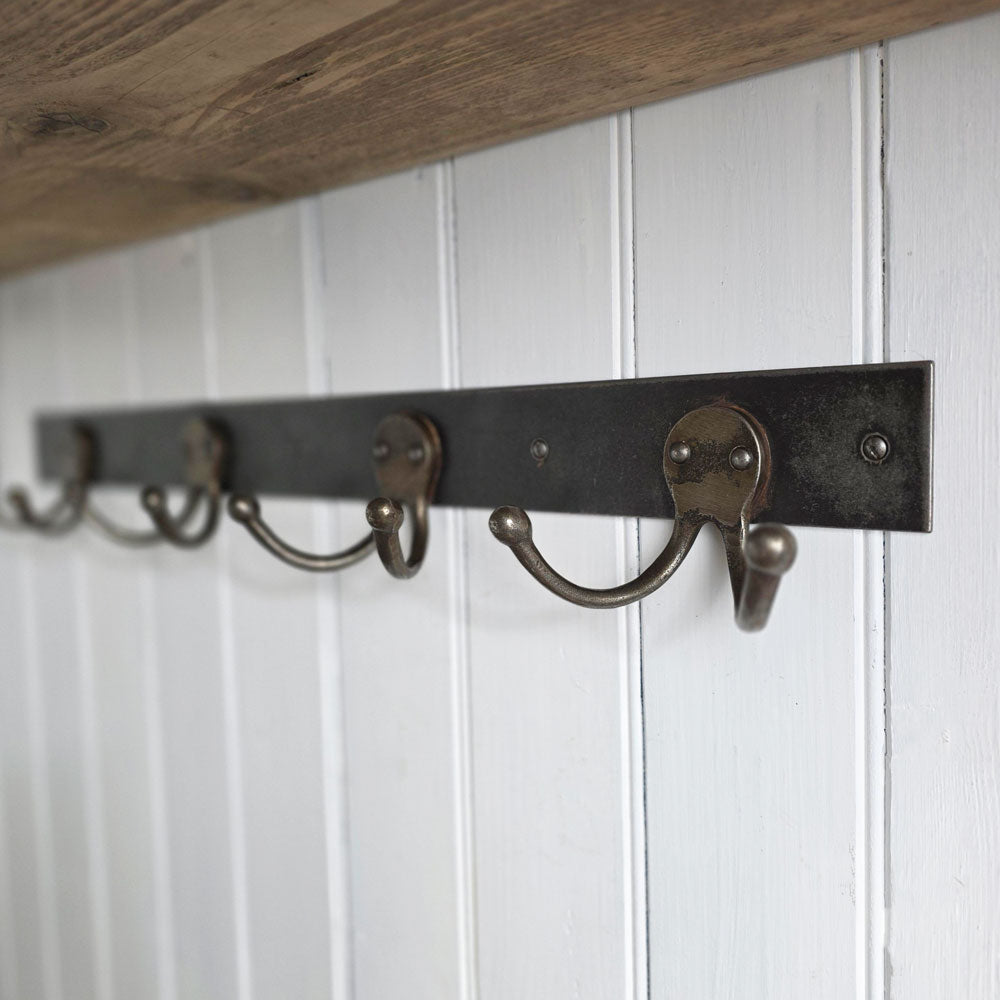 A rustic five-hook coat rack with a dark metal bar and bronze-toned hooks, mounted on a white paneled wall under a wooden shelf.