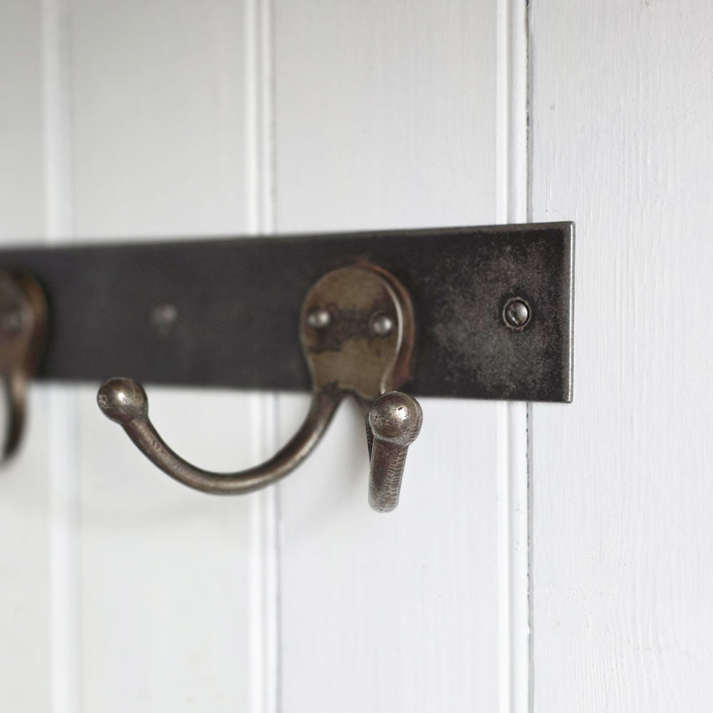 A close-up of a rustic metal coat rack with dark weathered horizontal bars and curved hooks mounted on a white paneled wall.