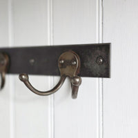 A close-up of a rustic metal coat rack with dark weathered horizontal bars and curved hooks mounted on a white paneled wall.