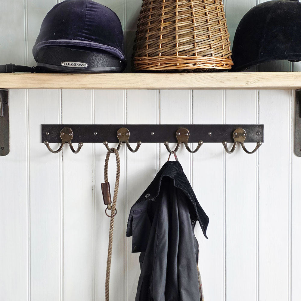 A rustic black metal wall-mounted coat rack with five hooks hanging in a white paneled wall. A dark coat hangs from the center hook, and a rope accessory hangs from the left hook.
