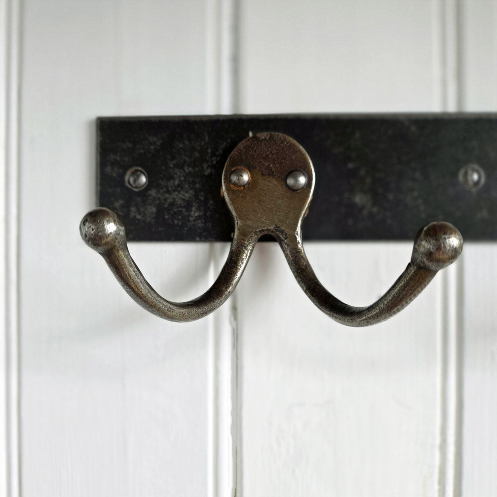 A rustic double hook coat rack made of weathered bronze metal, mounted on a black metal bar against a white paneled wall.