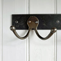 A rustic double hook coat rack made of weathered bronze metal, mounted on a black metal bar against a white paneled wall.