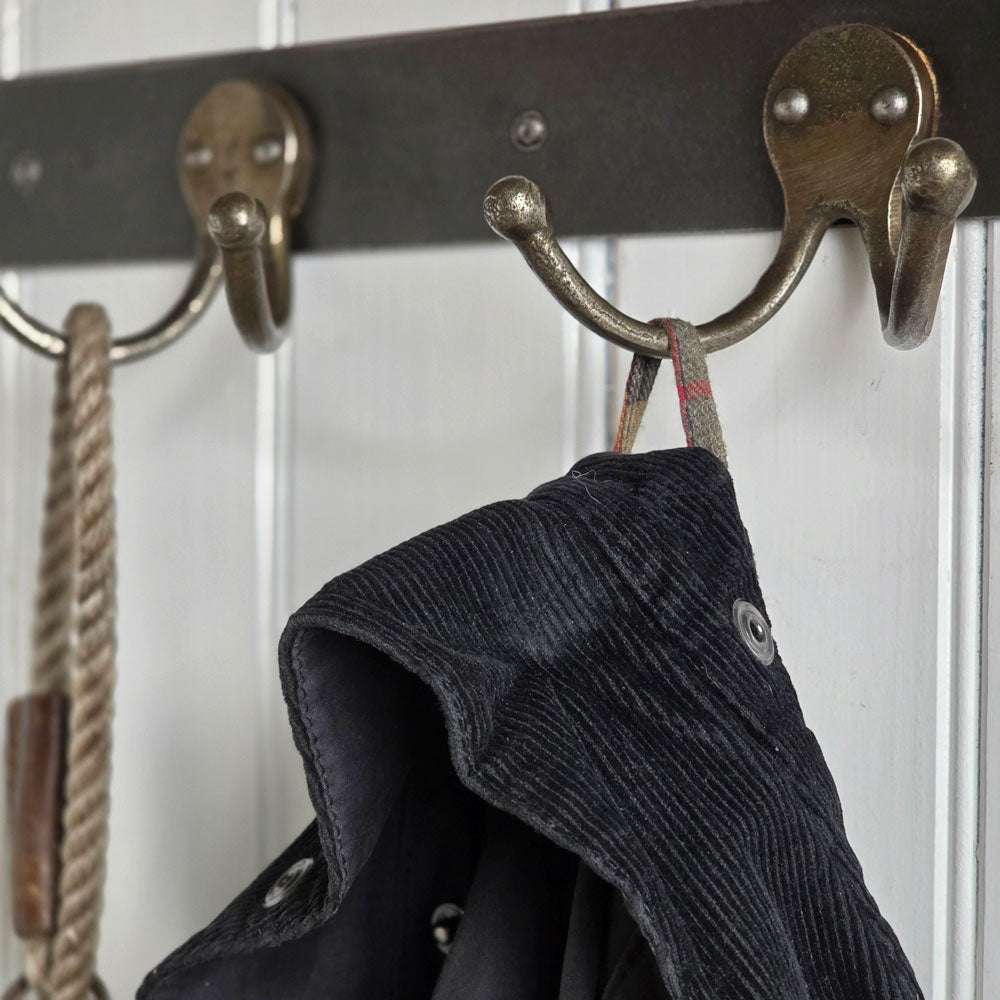 A close-up of rustic bronze metal coat hooks mounted on a dark horizontal bar, with a dark corduroy jacket hanging from one hook and a rope accessory hanging from another against a white paneled background.