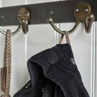 A close-up of rustic bronze metal coat hooks mounted on a dark horizontal bar, with a dark corduroy jacket hanging from one hook and a rope accessory hanging from another against a white paneled background.