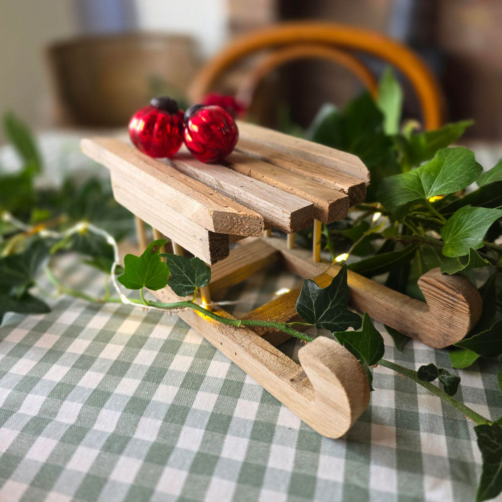 Miniature wooden sleigh on a green checked tablecloth with Christmas foliage and fairy lights.